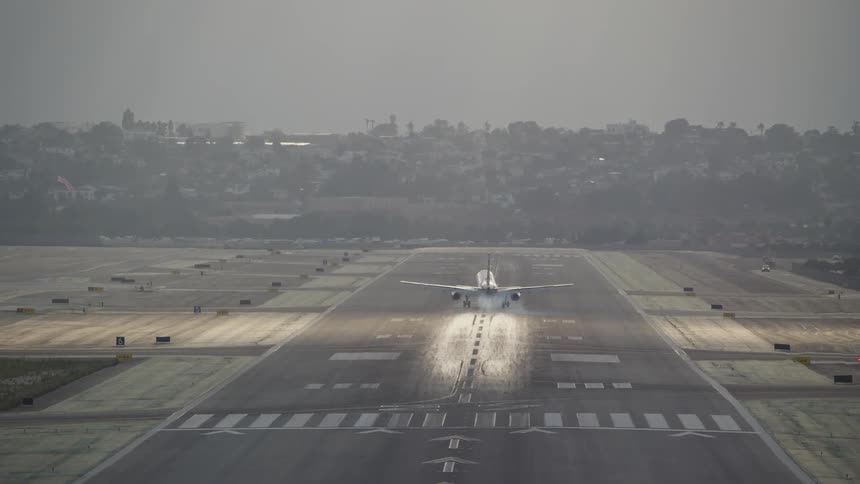 The video shows an airplane landing on a runway viewed from behind and at a distance the runway has visible markings and there are faint reflections of light on its surface as the plane descends