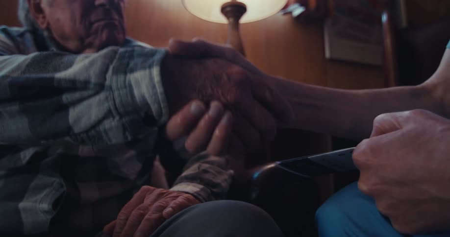 An older person in a plaid shirt shakes hands with someone in blue scrubs holding a clipboard sitting near a lamp and wooden paneling