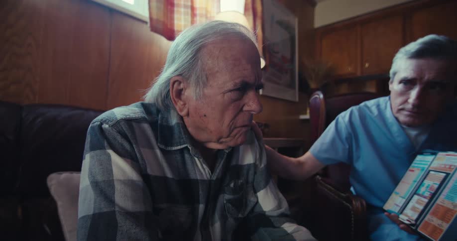 An elderly man sits on a couch as a healthcare worker in blue scrubs shows a colorful chart on a clipboard and rests a hand on his shoulder in a cozy woodpaneled room