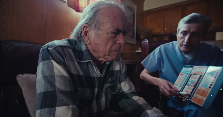 A male wearing scrubs is showing an elderly man a brochure while both men are sitting in a living room
