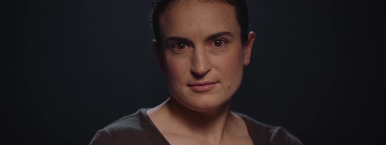 A woman with dark hair pulled back looks directly into the camera with a subtle smirk as light gradually illuminates her face against a dark background then slowly fades back into shadow