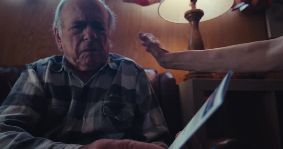 An elderly man sitting on a couch beside a lamp being comforted by someone after receiving a flyer