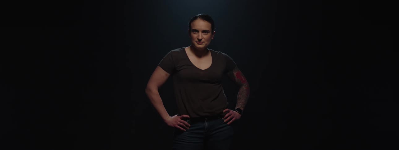 A young woman stands on a dark stage gradually illuminated by light with both hands resting on her waist