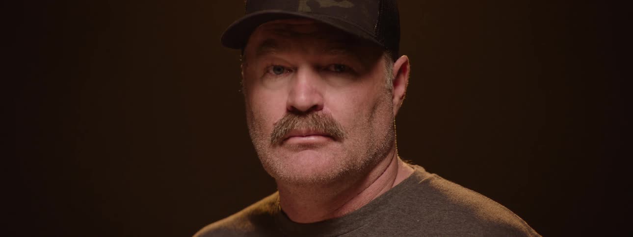 A middleaged man with a mustache wearing a hat has his face revealed by stage lights as he spins 360 degrees