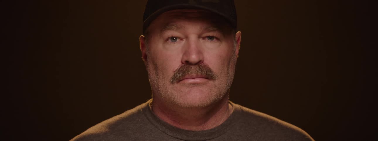 An officer on a stage turns 360 degrees to reveal his face wearing a baseball hat
