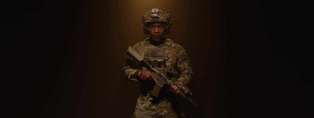 A man in a military uniform wearing a helmet and holding a weapon stands on a stage turning 360 degrees to reveal himself