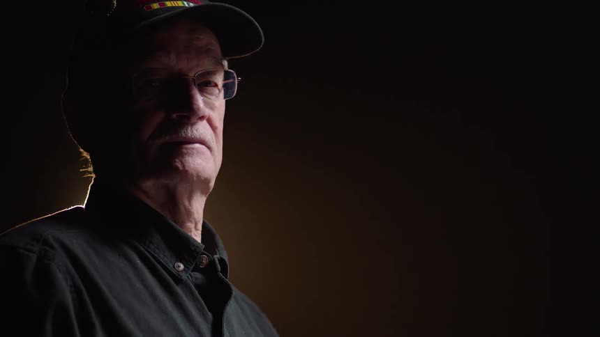 An older man wearing glasses and a cap with a mustache looks directly at the camera with a neutral expression before turning his head to the right