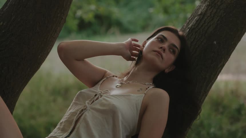 A young woman lying on a tree trunk branch in the woods stroking her hair and lost in thought