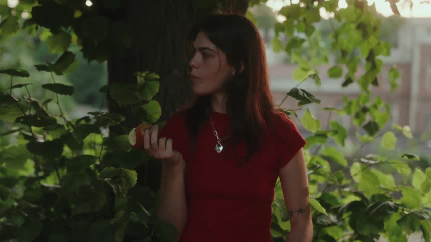 Young woman eats a fruit while standing under tree outdoors during daytime
