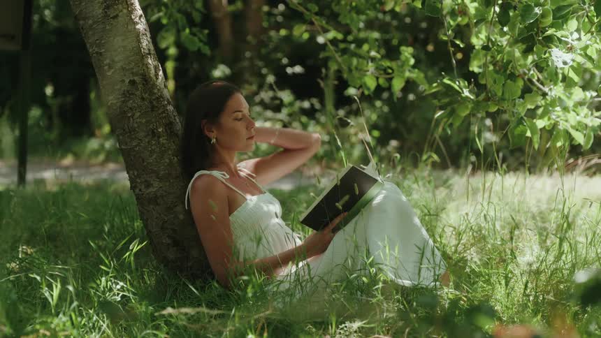 A young woman resting against a tree sitting on the grassland floor outdoors reading a book