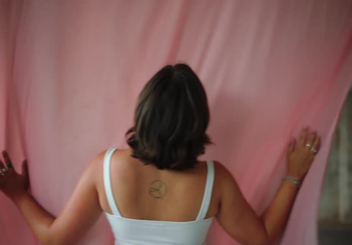 Woman with a tattoo on her upper back sways next to a fluttering piece of fabric while wearing multiple rings