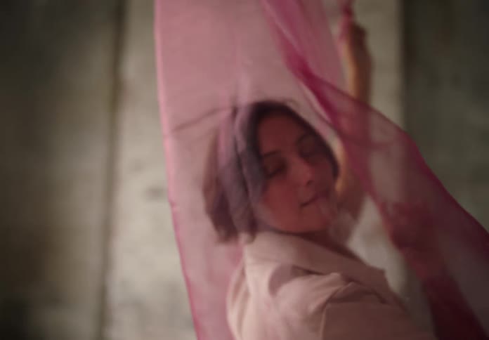 A woman poses gracefully with her arm raised behind a flowing pink fabric