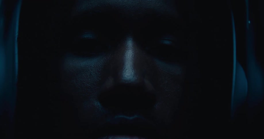 The video shows an extremely closeup and dark shot of a persons face with minimal detail visible except for the faint outlines of their features theres a subtle blue light casting a glow possibly from a screen creating a somber