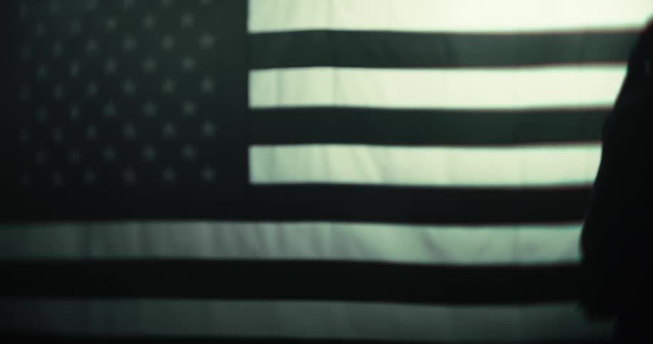 A black person with long braids walks in front of a large blackandwhite american flag partially in shadow