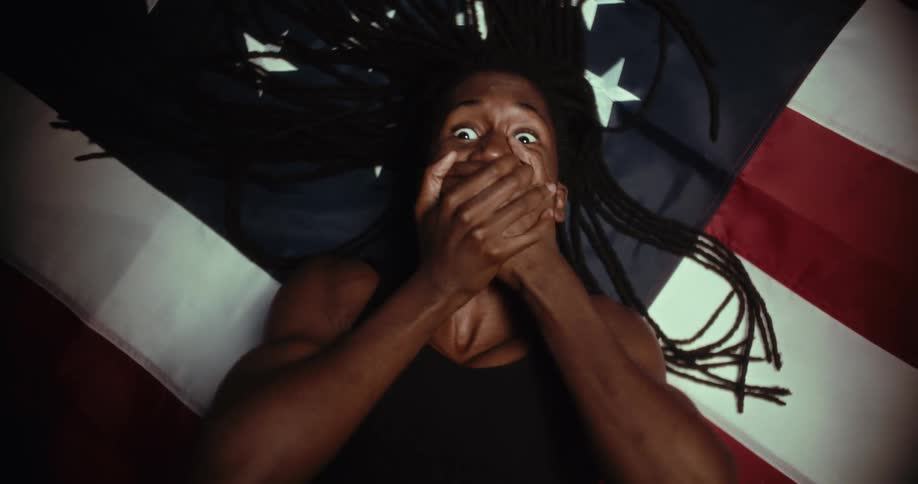 The video shows a person with dreadlocks lying on an american flag looking up at the camera with wide surprised eyes their hands are covering their mouth conveying an expression of shock or speechlessness