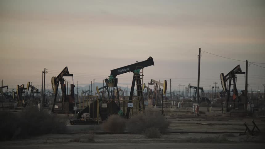 Multiple oil pump jacks operate across a dry dusty landscape under a hazy sky forming a dense field of industrial activity at dusk
