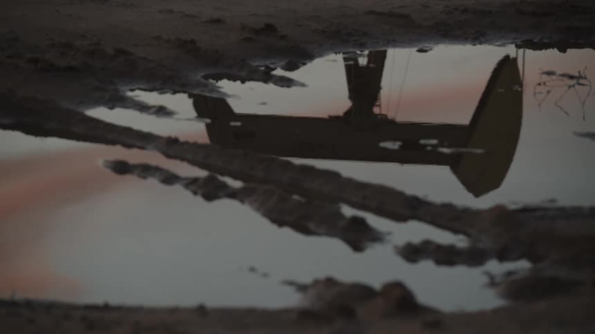 An oil pump jack is reflected in a muddy puddle at dusk with soft pink and blue hues in the water hinting at a fading sunset
