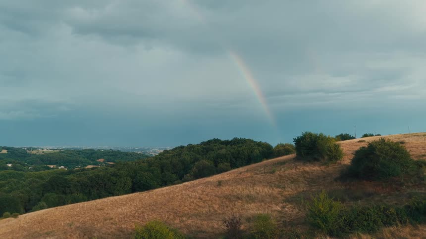 The video shows a scenic overview of a vast hilly landscape under a dramatic sky with a visible rainbow arcing across the horizon the foreground features a dry grassy hill while the background reveals dense green forests and distant mountains or city