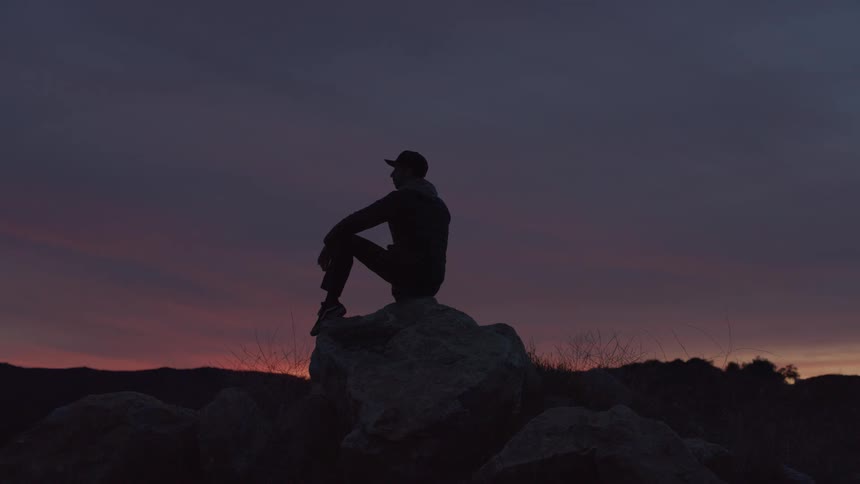 Silhouette of a man in a cap sitting on a rock in an outdoor setting at dawn