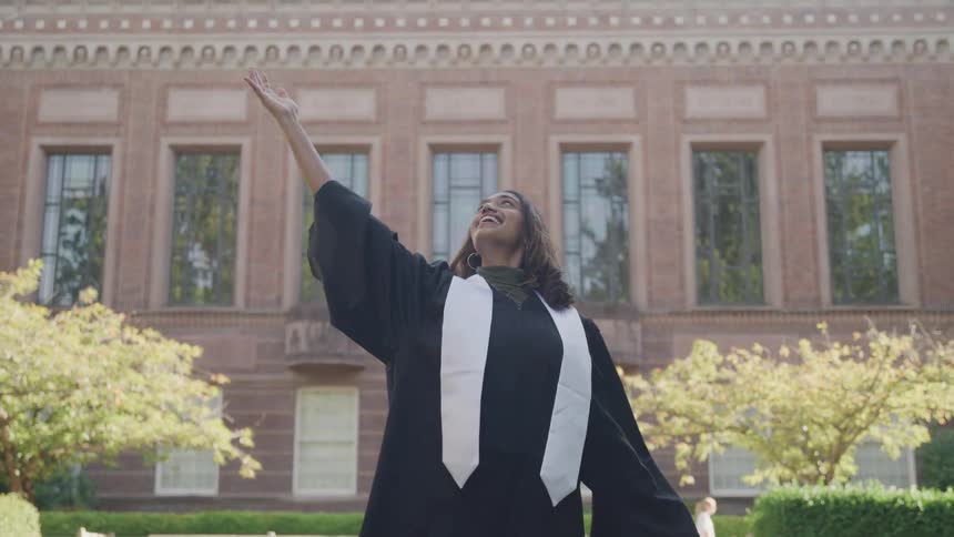 The video features a female graduate in a black cap and gown standing outdoors in front of a large brick building she joyfully removes her cap and smiles broadly celebrating her accomplishment