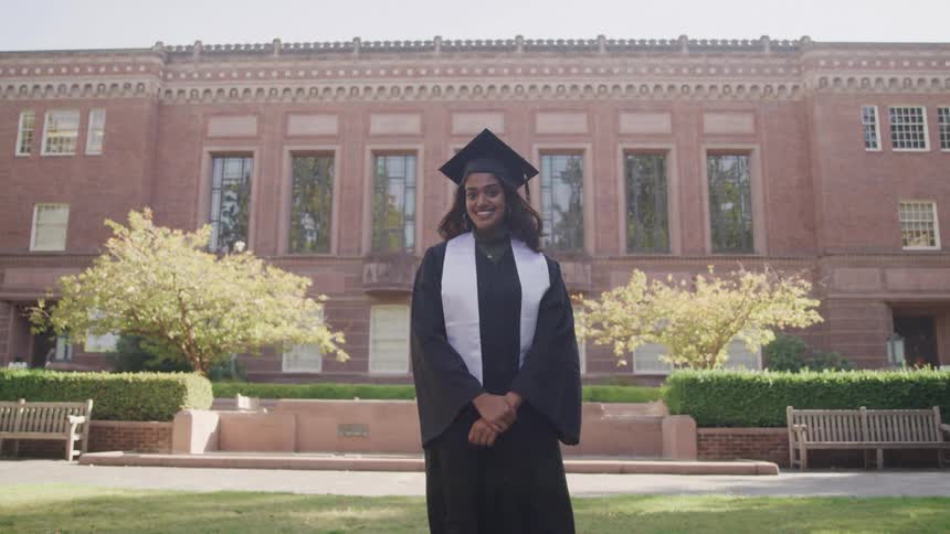 The video features a female graduate in a black cap and gown standing outdoors in front of a grand brick university building with many windows she stands proudly facing the camera with a confident and happy expression