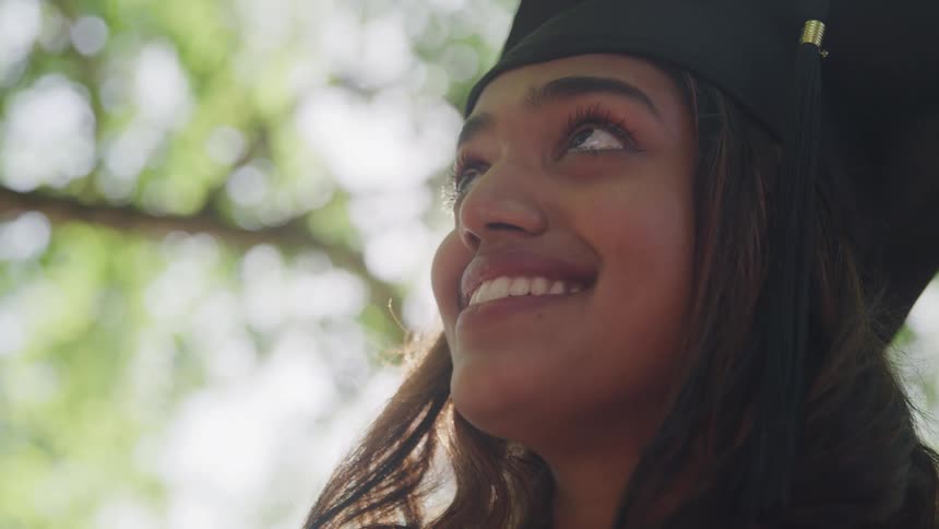 The video features a female graduate in a black cap and gown looking upwards with a joyful expression her eyes wide and a smile on her face bright sunlight streams from the upper left creating lens flares and a hopeful celebratory atmosphere
