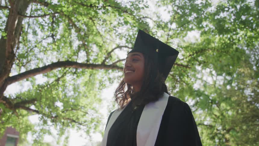 Lowangle shot of a person dressed in a black graduation cap and gown with a white stole looking upwards with a warm smile
