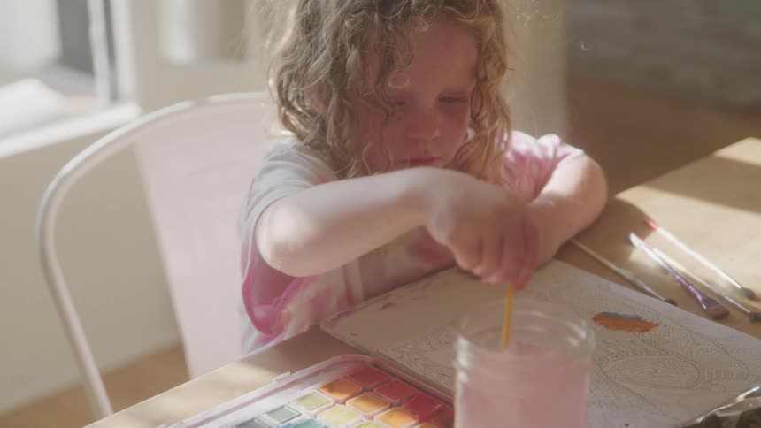 The video shows a young child with curly lightcolored hair sitting at a table and focused on painting they are dipping a paintbrush into a glass of pink water and then applying paint to a coloring book indicating a creative and engaging activity