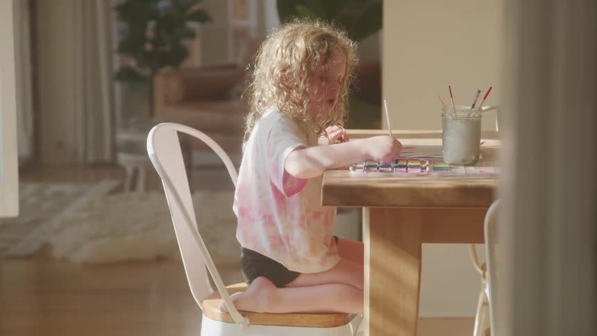 Young child with curly blonde or light brown hair sitting at a wooden table engaged in painting with watercolors