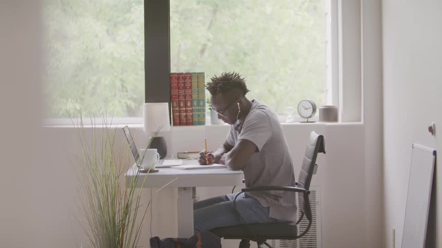 The video shows a male student with dreadlocks wearing earbuds sitting at a white desk and diligently writing in a notebook while looking at a laptop the setting appears to be a home office or study area with a window and a bookshelf visible in the ba