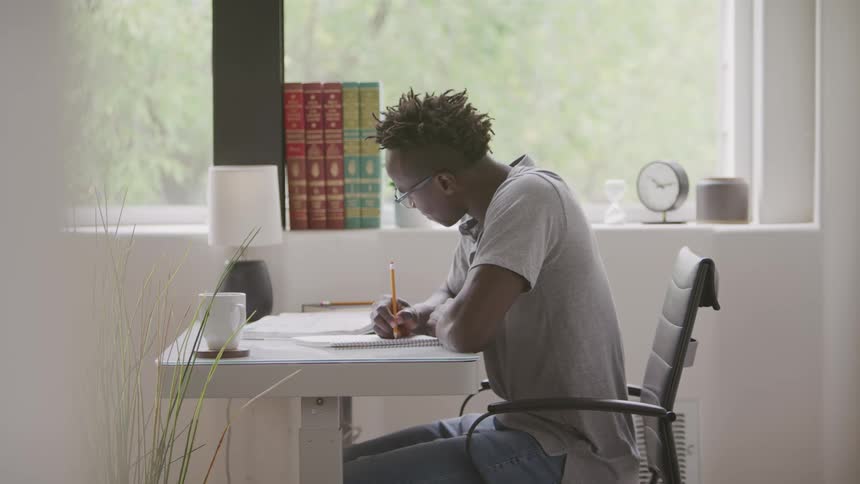 The video shows a male student with dreadlocks wearing glasses sitting at a desk and diligently writing in a notebook or textbook with a pencil the setting appears to be a welllit room with a window providing natural light and a bookshelf filled with