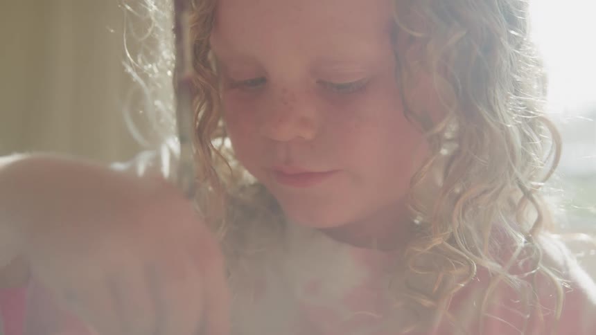 The video features a young child with curly lightcolored hair intently focused on an activity they are seen dipping a paintbrush into a glass of pink water before transferring it to a coloring book indicating they are engaged in painting