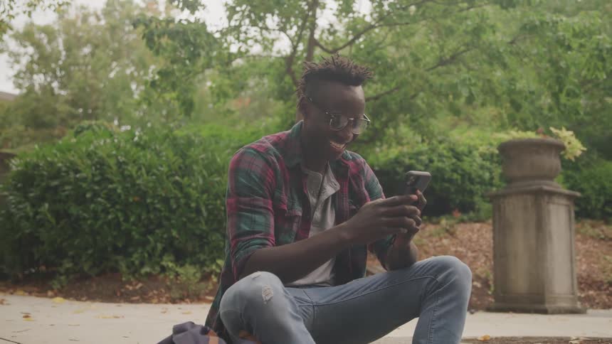 The video features a man with dreadlocks wearing glasses and a plaid shirt actively engaged with his phone while sitting outdoors in a parklike setting the background includes lush greenery and a decorative stone urn adding to the serene atmosphere