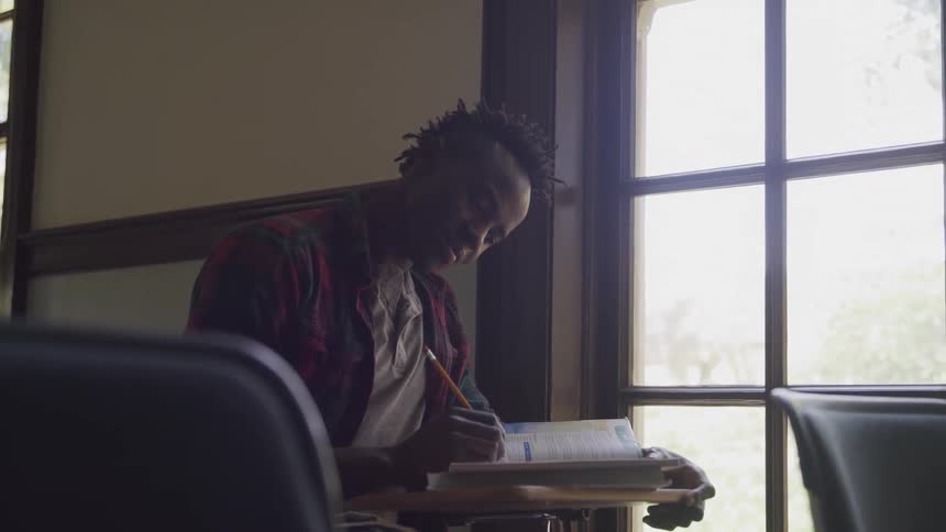 The video shows a male student with dreadlocks wearing a plaid shirt sitting at a desk by a large window he is attentively writing in a textbook with a pencil suggesting a focused study session