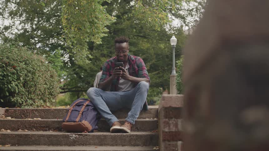Man sitting on the steps in a park smiles and texts on his smartphone an autumn day