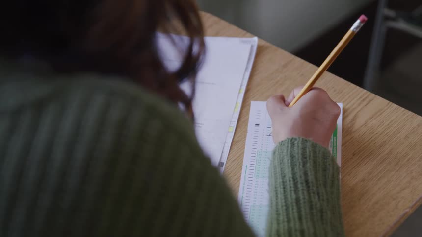 The video shows a closeup overtheshoulder view of a person with long dark hair wearing a green sweater meticulously filling in a multiplechoice bubble sheet with a pencil on a wooden desk papers are scattered around the work surface suggesting an