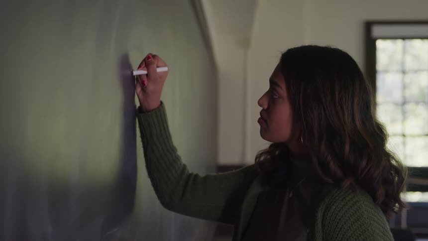 Woman with a white chalk writing on a blackboard in the classroom