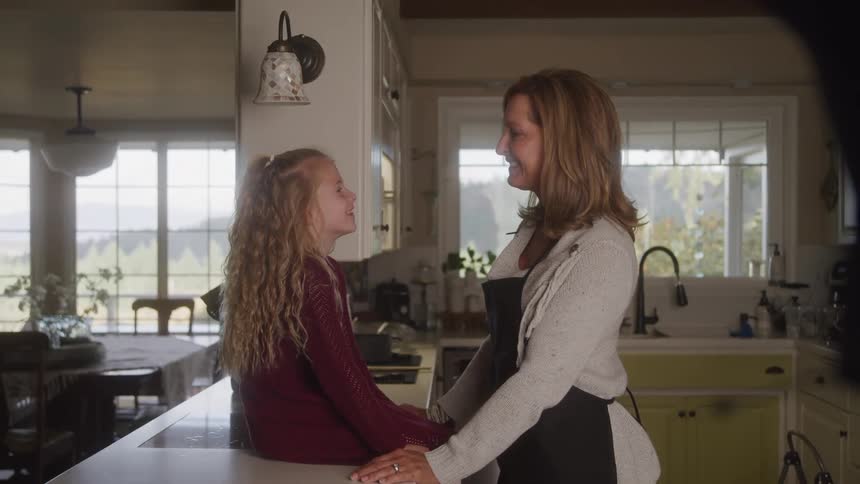 The video features an adult woman and a young girl with long curly blonde hair standing in a modern kitchen both looking at each other and smiling they are holding hands indicating a warm and affectionate interaction