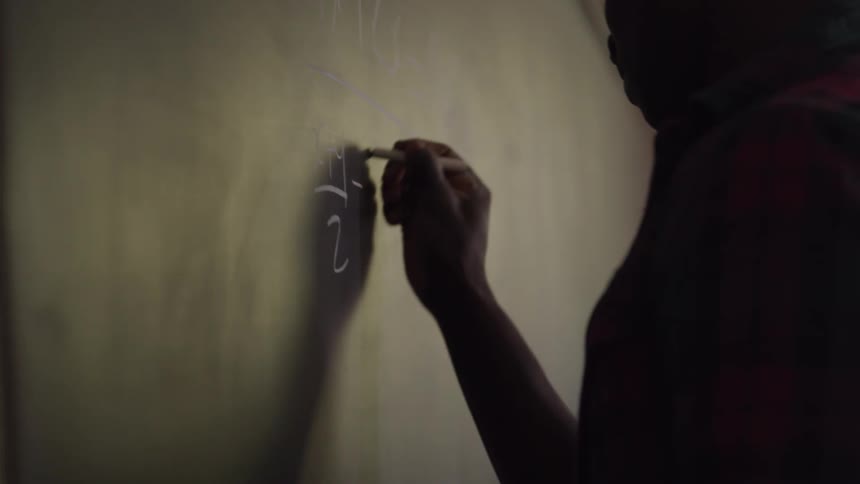 The video shows a persons hand holding a piece of chalk and writing on a dark surface possibly a chalkboard the scene is dimly lit with the persons profile silhouetted against the background