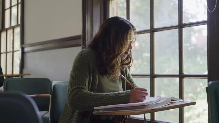 The video shows a female student with long dark hair wearing a green cardigan diligently writing in a notebook in a classroom natural light streams in from a large window in the background