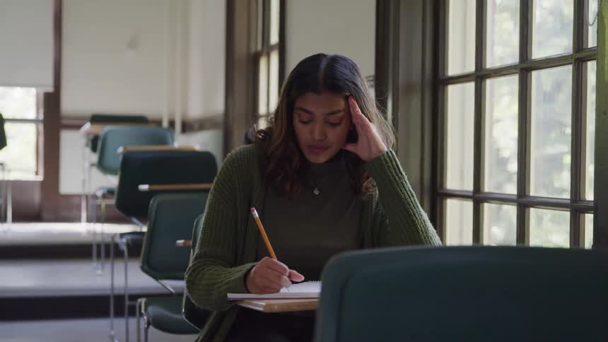 A young woman is in a classroom alone writing on paper while sitting at a desk