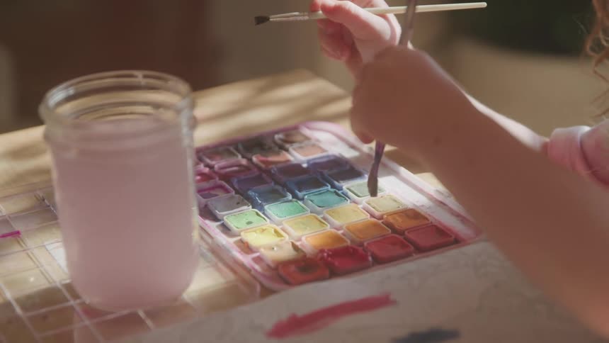 The video shows a closeup of a childs hands dipping a paintbrush into a colorful watercolor palette and then applying paint to paper with a jar of water nearby
