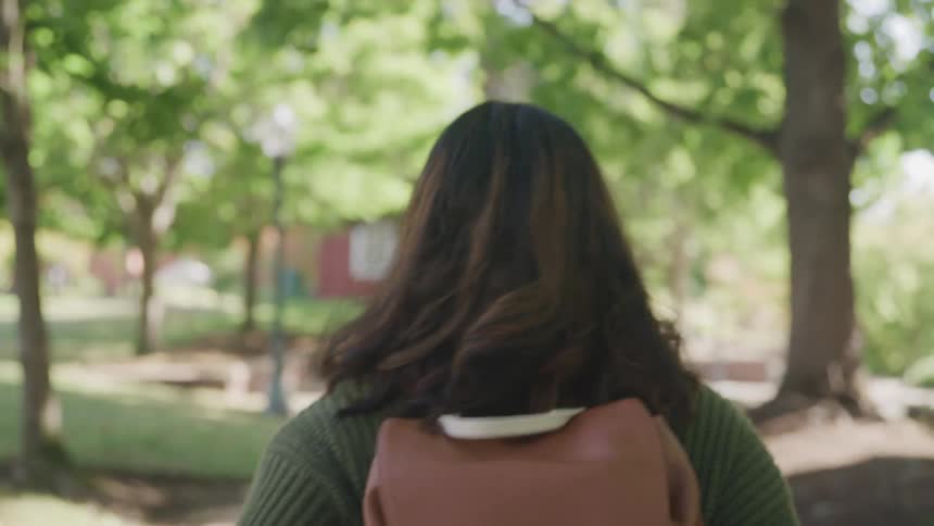 The video shows a student walking away from the camera on a college campus wearing a brown backpack and a green sweater with large academic buildings visible in the background