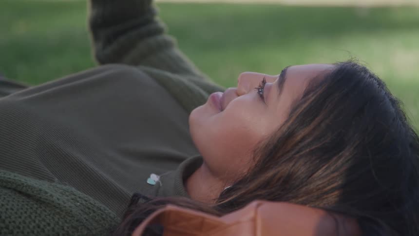 The video shows a closeup of a person lying down on a surface looking upwards with a contemplative expression the person has long dark hair and is wearing a green top suggesting a relaxed outdoor setting such as a park or garden