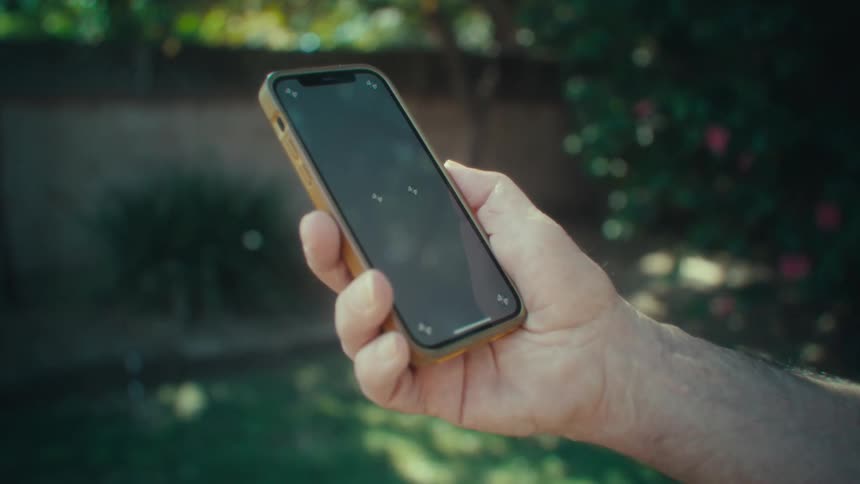 The video shows a person holding a smartphone outdoors with the focus on the hand and phone as it is presented against a blurry green background the phone has a lightcolored case and a dark blank screen