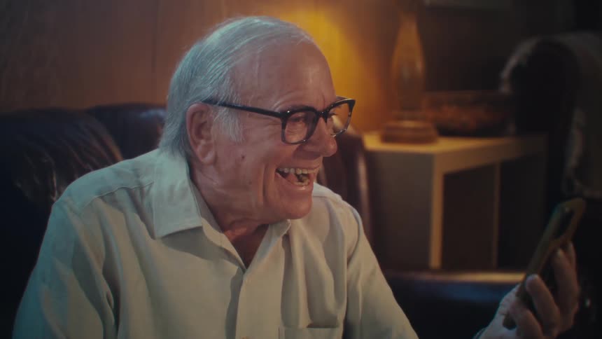 Elderly man sitting on a sofa laughing while holding a smartphone in front of him