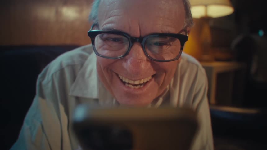 Elderly man with glasses sitting on a couch laughing to a smartphone thats in his hands
