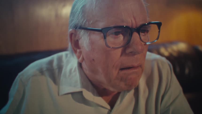 The video shows a side profile of an elderly man wearing glasses sitting in a dimly lit room looking at a smartphone the room appears to have traditional wooden paneling on the walls suggesting a cozy or vintage interior