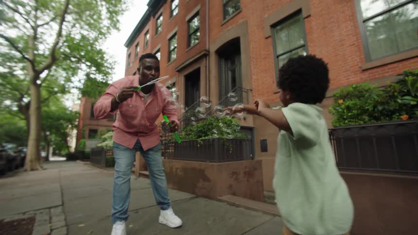 The video shows an adult and a young child on a city sidewalk with the adult creating bubbles using a bubble wand the child is actively engaged trying to catch the bubbles as they float by