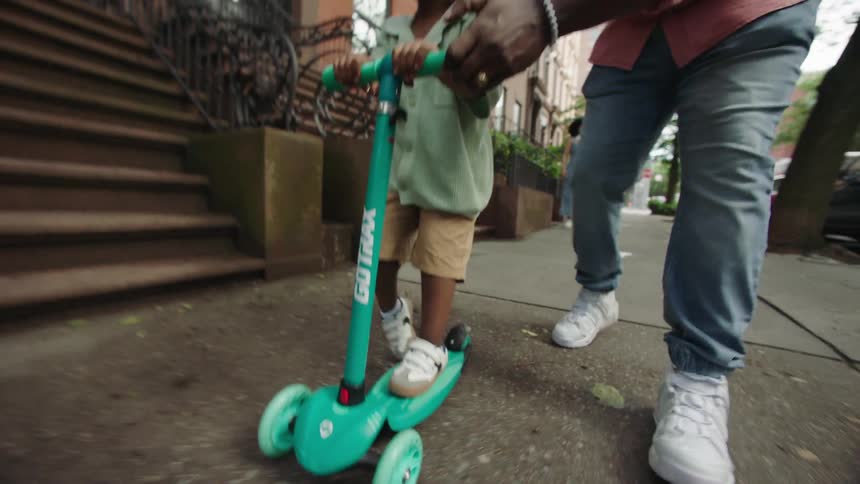 Father helps son ride scooter on sidewalk as mother stand behind